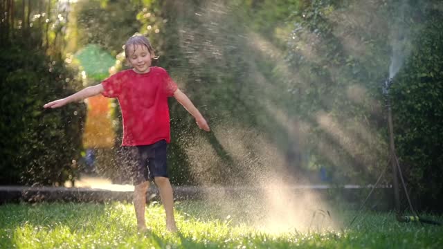 Slow motion video of funny little boy playing with garden sprinkler in sunny backyard.Elementary school child laughing, jumping and having fun with spray of water