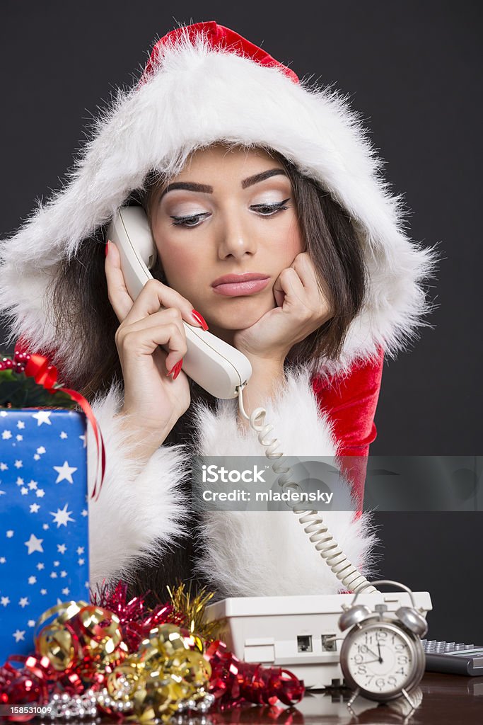 Unhappy Santa girl on the phone Portrait of unhappy Santa girl speaking on the phone with alarm clock, gift box and decorations on desk. Adult Stock Photo