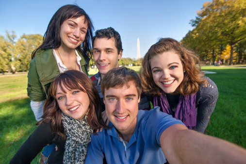 Group of five young men and women pose in front of the Washington Monument. Focus on the man up front holding the camera out and turning it on themselves.