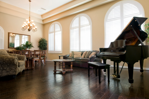 Upscale living room with wood floors, crown moulding and a grand piano. A dining area can be seen in the background with a high end chandelier.