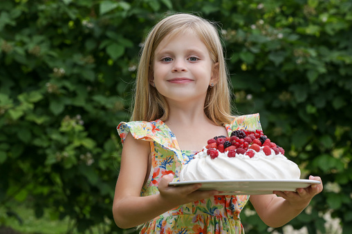 Prtrait of adorable little girl in floral summer dress holding tray with Pavlova cake