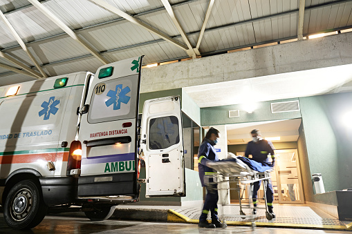 Full length view with blurred motion as uniformed man and woman in 20s and 50s wheel gurney into emergency room.