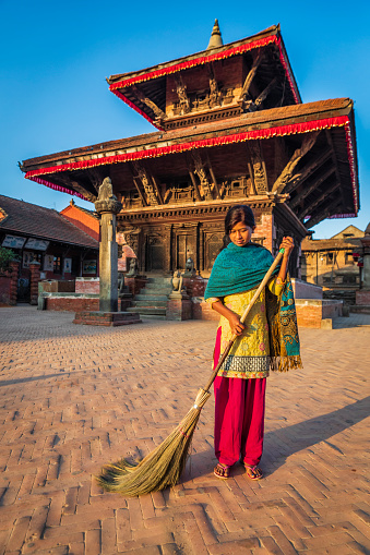 Nepali young woman sweeping on a d Durbar Square in Bhaktapur. An ancient Hindu temple on the background. Bhaktapur is an ancient town in the Kathmandu Valley and is listed as a World Heritage Site by UNESCO for its rich culture, temples, and wood, metal and stone artwork.