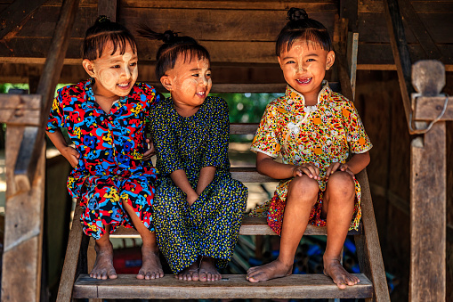Little Burmese girls with thanaka on their faces and an ancient hairstyle sitting next to their house in the village near Bagan, Myanmar (Burma)