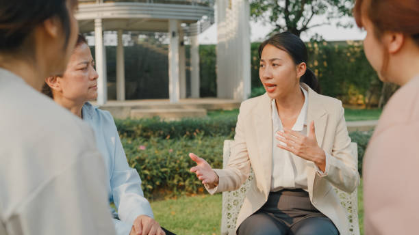 istock Group therapy session with diverse people sharing their sad problems and stories in park. Asian people sitting in circle at group therapy session with psychologist. Mental health. 1585442678