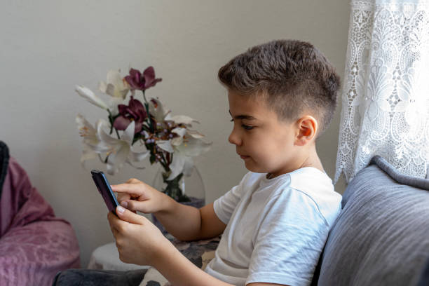 istock Little boy sitting in sofa and playing with cellphone at home 1585455497