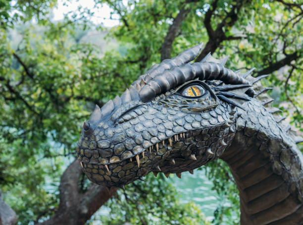 istock Fantasy art portrait real dragon head close-up sharp teeth beautiful eyes muzzle in dark gray-green scales, spikes. Creative big dinosaur toy of Jurassic period. Background summer nature green trees 1585471867