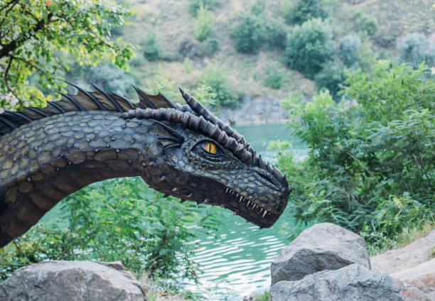 istock Fantasy art portrait real dragon head close-up sharp teeth beautiful eyes muzzle in dark gray-green scales, spikes. Creative big dinosaur toy of Jurassic period. Background summer nature green trees 1585471952