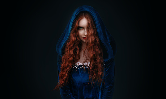 Portrait fantasy gothic red-haired woman witch. Vampire girl in blue medieval dress, vintage old historical style hood on head. Black background. Red lips hair flying soar in wind. halloween costume