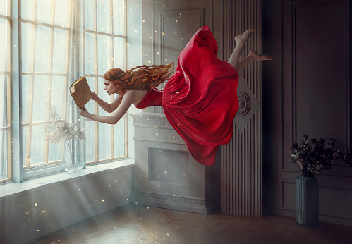Fantasy redhead woman soars floats flies in air. Art photo levitation. Girl fairy princess reads magic book, divine light from window. Red midi dress, lon hair flutters in wind. Room classic interior.