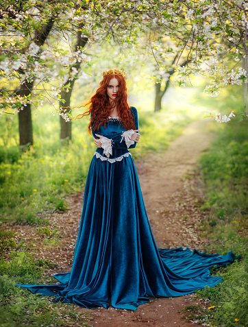 Redhead fantasy woman queen. Blue long velvet medieval dress, vintage clothing. Red curly hair flying waving in wind. Summer nature green flowering trees garden footpath path. Girl Princess walking.