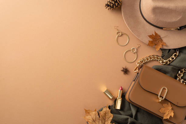 Classic female attire with autumnal touch. Top view of brimmed hat, grey scarf, handbag, gold earrings, lip color, scattered leaves, anise, pine cone on beige backdrop with blank area for text or ad vestimenta femenina clásica con toque otoñal. vista superior de sombrero de ala ancha, bufanda gris, bolso, pendientes dorados, color de labios, hojas dispersas, anís, cono de pino sobre fondo beige con área en blanco para texto o anuncio