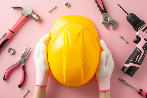 First person top-down view of female worker's hands, adorned in work gloves, holding safety helmet. Tools like screwdriver, wrench, drill, pliers, screws, nuts and hammer lie beside pink background