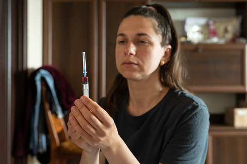 The nurse is holding a syringe with medicine. The red liquid in the syringe is a vitamin of group B. The nurse is preparing for a medical procedure. The nurse works at the patient's home.