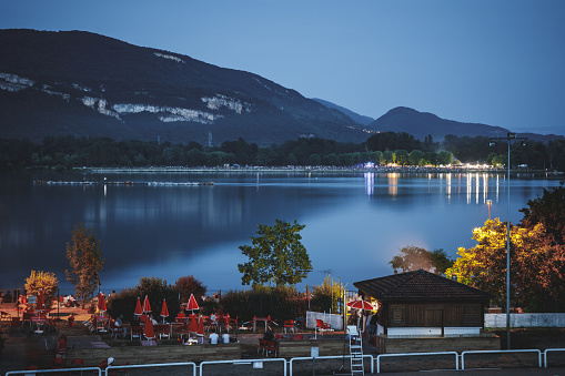 Horizontal dark photography at twilight of the Rhone riverbank, with small snack bar and unrecognizable person, and mountain reflections in water. This photo was taken in Montalieu-Vercieu city, in Isere, Auvergne-Rhone-Alpes region in France, Europe.