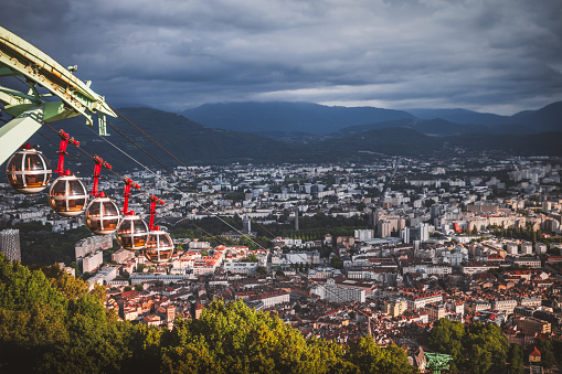 Majestic panorama over Grenoble city at sunset, with bright sunlight through clouds during a summer day, and with famous cable car on left. Taken in the department of Isere, Auvergne-Rhône-Alpes region in France, Europe.