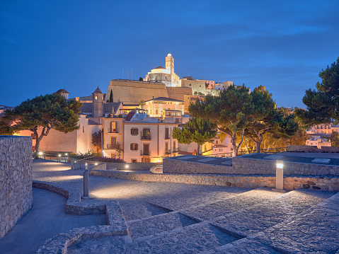 Wide-angle view of Dalt Vila, the old town centre of Ibiza, and its renowned iconic skyline dominated by the cathedral church of Santa Maria de les Neus, at nightfall. The intense blue sky of a Mediterranean summer early night, scenic street and building lighting, a long flight of paved steps, lush pine trees. High level of detail, natural rendition, realistic feel. Developed from RAW.