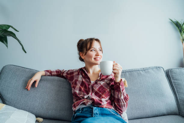 istock Young smiling woman sitting on sofa and looking side up while drinking coffee or tea. Young brunette woman relaxing after housekeeping, home cleaning. Portrait of relaxed female resting at home 1585575207