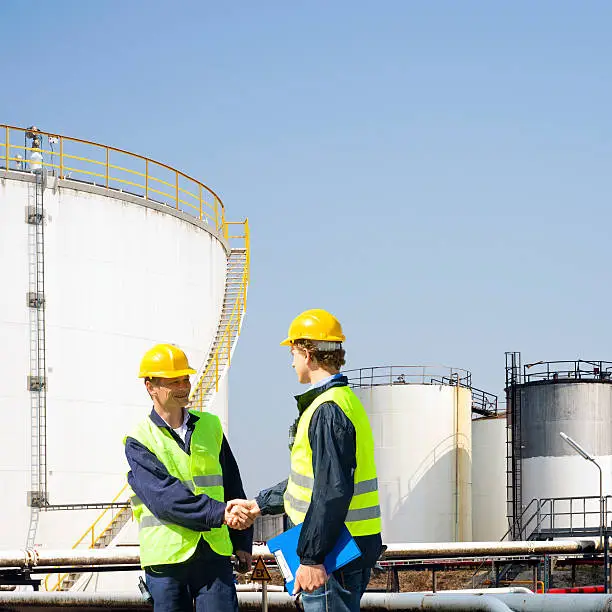 Two oil industry workers shaking hands in front of the storage tanks of a petrochemical refinary