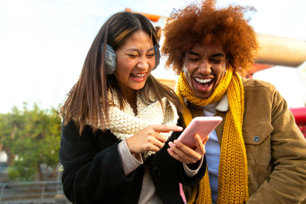 Multiracial couple having fun, laughing using phone outdoors on winter day. Friends watching online videos using phone.