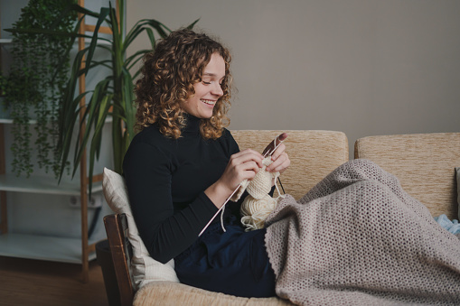 Side view portrait of smiling woman knitting using ribbon yarn and crochet needles. Happy creative female posing at home comfortable interior enjoy art work or hobby.