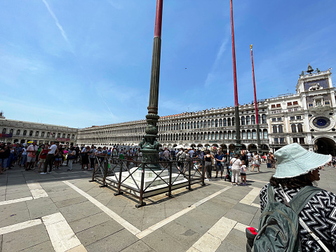 St Mark's Square, Venice, Italy - July, 12 2023: Stock photo showing close-up view of tourists milling around three flagpoles in St Mark's Square (Piazza San Marco) and walking past the Doge's Palace (Palazzo Ducale). These flagpoles were once ship’s masts from which the flags of the Republic of Venice, Italy and European are flown.