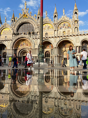 St Mark's Square, Venice, Italy - July, 12 2023: Stock photo showing close-up view of tourists walking past puddles in St Mark's Square (Piazza San Marco) past Basilica di San Marco (St Mark's Cathedral). The puddles are caused by flooding due to strong winds and high tides pushing Adriatic Sea water into the Venetian Lagoon, a phenomenon called acqua alta (high water).