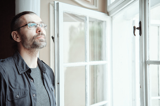 Bearded man in glasses stands at an open window and looks thoughtfully into the distance.