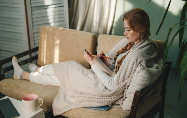 istock Young minded woman writing down memories in notebook diary sitting on sofa relaxing her spend free spare time in living room indoor. Creativity, art and education concept. 1585835755