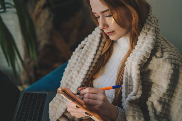 istock Young woman wrapped in plaid writing down memories in notebook diary sitting on sofa spending free spare time in living room. People lounge concept 1585836300
