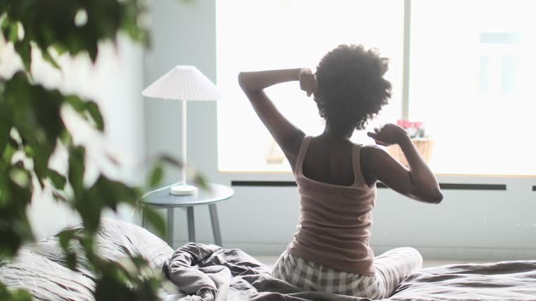 Young adult black woman waking up and stretching in bed in the morning in modern bedroom