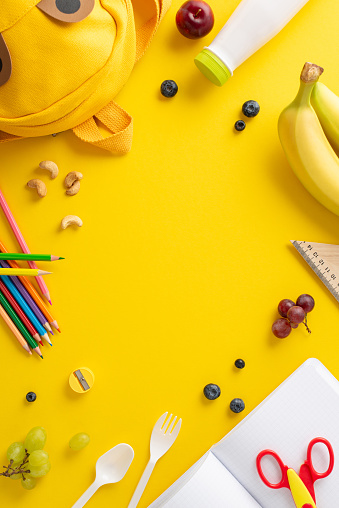 Instill wellness values at school - vertical top view fresh fruits, berries, nuts, yogurt, cutlery, stationery, color pencils, scissors, diary, funny backpack on yellow background with space for text