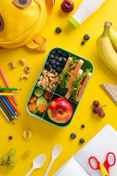 Elevate school meal experience - vertical top view of lunchbox filled with nutritious bites, cutlery, yogurt, stationery, color pencils, copybook, scissors, funny schoolbag on yellow backdrop