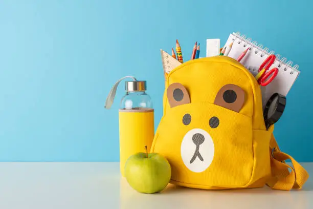 Photo of School days in one frame: side view photo of neatly arranged desk with school supplies, water bottle, apple, cute teddy-bear schoolbag with stationery against a blue wall background with space for ad