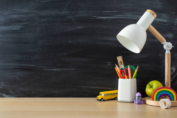 istock Enhance your study sessions with this student's workplace. Side view photo featuring desk lamp, stationary and toys on wooden table on blackboard isolated background, perfect for promotional messages 1585995411