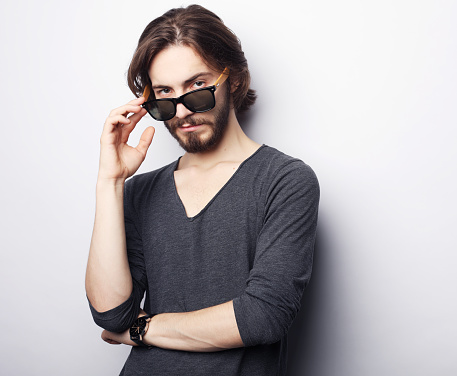 Handsome young male model, with beard and mustache, wearing sunglasses, stylishly dressed. Portrait on a gray background.