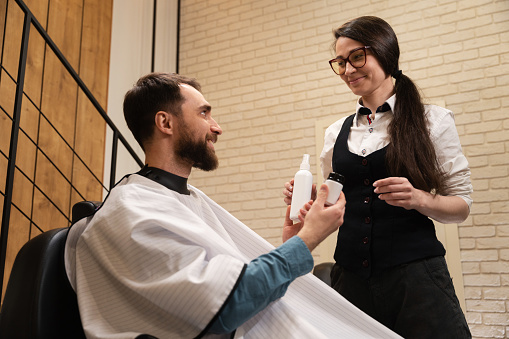Young woman in a barbershop demonstrates male care products, the client is seated in a comfortable chair