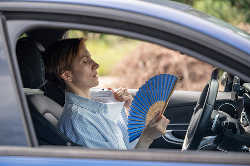Tired exhausted middle aged woman waving blue fan suffers from stuffiness driving car on summer hot weather. Overheating, sultriness, high temperature, swelter in car with broken air conditioner.