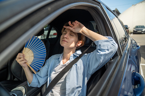 Exhausted tired middle aged woman drives car waves blue fan suffers from stuffiness stands in urban traffic jam in summer hot weather. Overheating, high temperature in car with broken air conditioner.