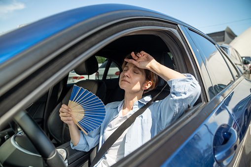 Exhausted tired middle aged woman drives car waves blue fan suffers from stuffiness stands in urban traffic jam in summer hot weather. Overheating, high temperature in car with broken air conditioner.
