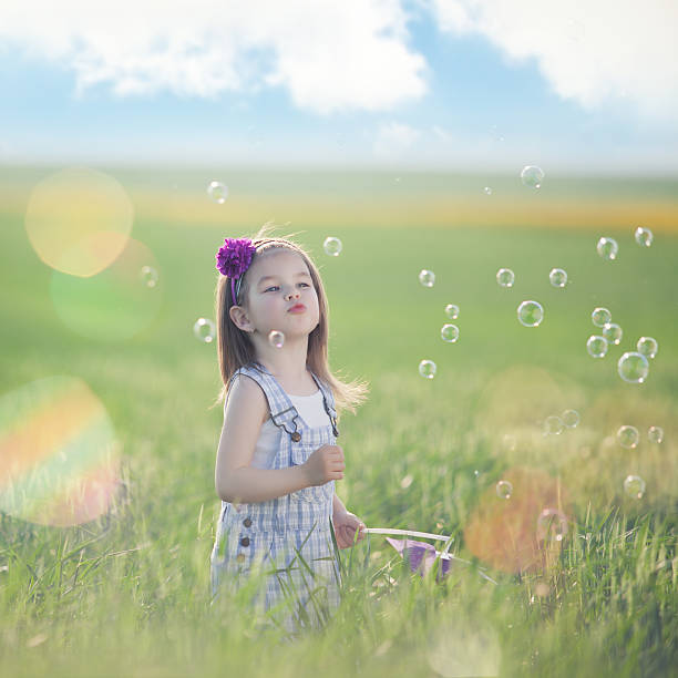 Little girl playing on a wheat field with a pin-wheel surrounded by soap bubbles  