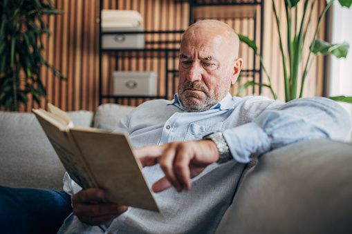 Senior man reading a book sitting on sofa at home.