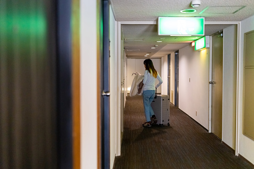 Asian woman tourist walking down hotel corridor with suitcase to her room. Businesswoman working outside office. Attractive girl traveler enjoy travel on holiday vacation. Hotel and resort business concept.