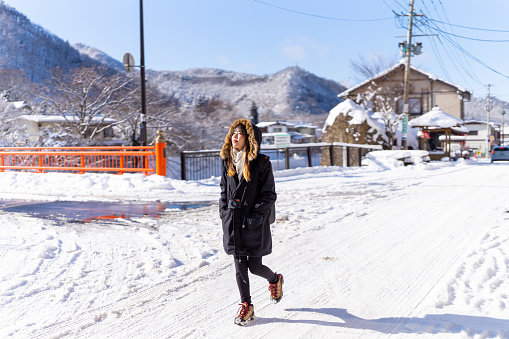 Young Asian woman in winter coat walking down street in small town covered in snow in snowing day. Attractive girl traveler travel local village near the mountain in Japan on winter holiday vacation.