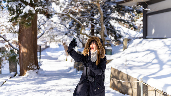 Asian woman in winter coat using mobile phone taking selfie during travel on forest mountain in snow day. Attractive girl travel local village covered in snow in Japan on winter holiday vacation.