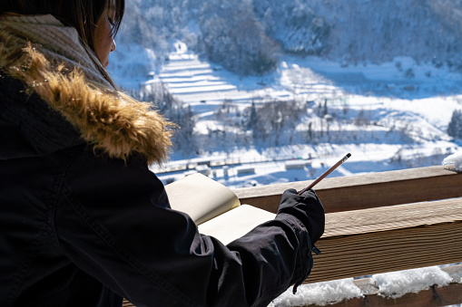 Asian woman writing on diary book at wooden seat pavilion rest stop on the mountain during travel on road trip in Japan. Attractive girl enjoy outdoor lifestyle travel on winter snow holiday vacation.