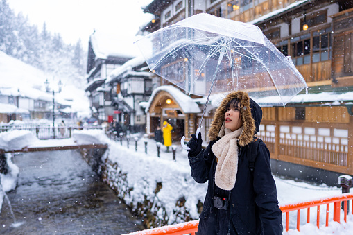 Young Asian woman tourist holding umbrella walking down footpath at onsen area Ginzan Onsen in Yamagata prefecture, Japan in snowy day. Attractive girl travel local village landmark on winter vacation.