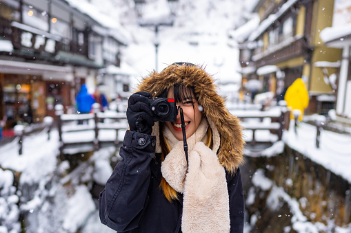 Young Asian woman tourist using digital camera taking picture during travel Ginzan onsen area in Yamagata prefecture, Japan in snow day. Attractive girl travel local village landmark on winter vacation.