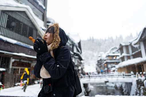 Young Asian woman tourist in winter coat eating traditional Japanese street food during travel at Ginzan onsen area in Yamagata, Japan in snow day. Attractive girl travel landmark village on winter vacation.