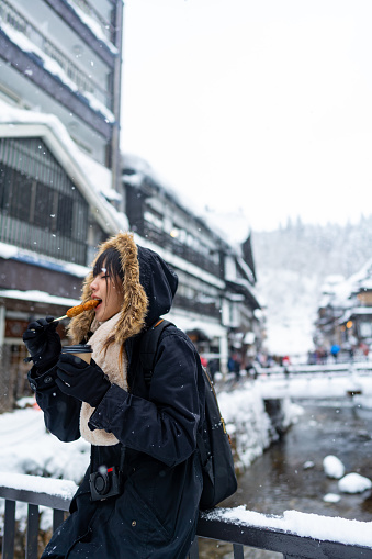 Young Asian woman tourist in winter coat eating traditional Japanese street food during travel at Ginzan onsen area in Yamagata, Japan in snow day. Attractive girl travel landmark village on winter vacation.